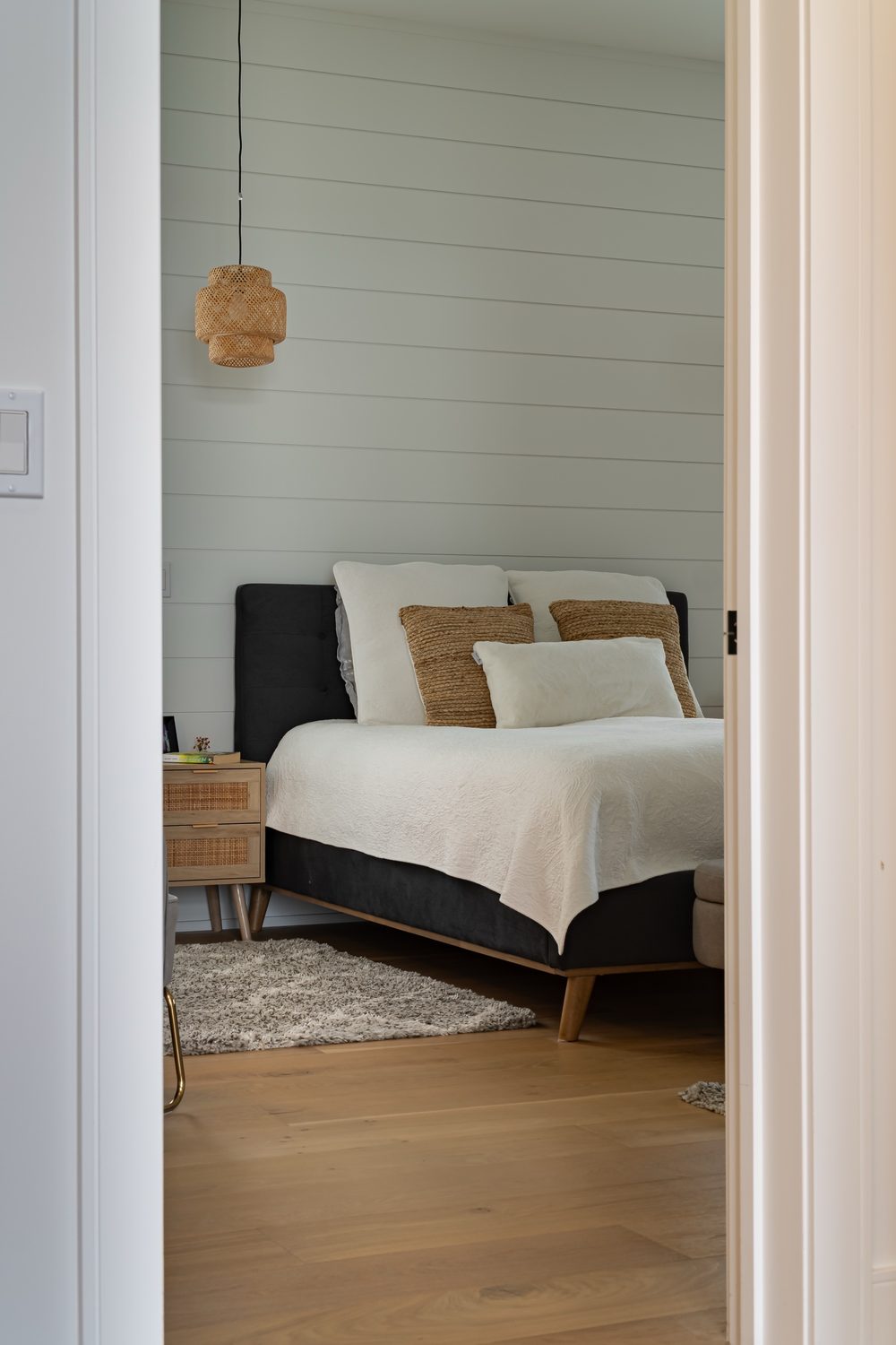 View through a doorway into a bedroom with a shiplap wall, a woven pendant light and a dark upholstered bed