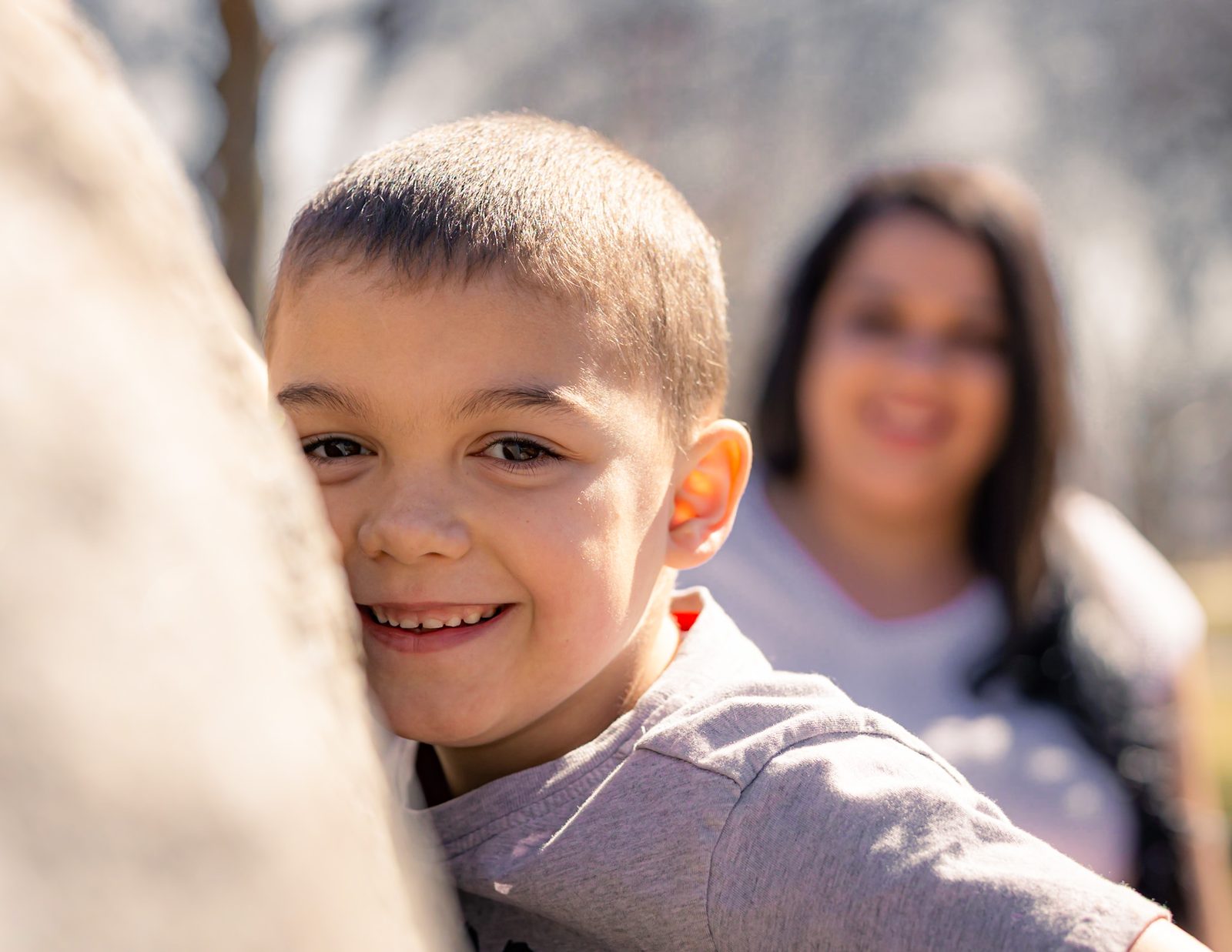 Smiling boy peeking from behind a soft foreground in bright sunlight, his mother out of focus behind him