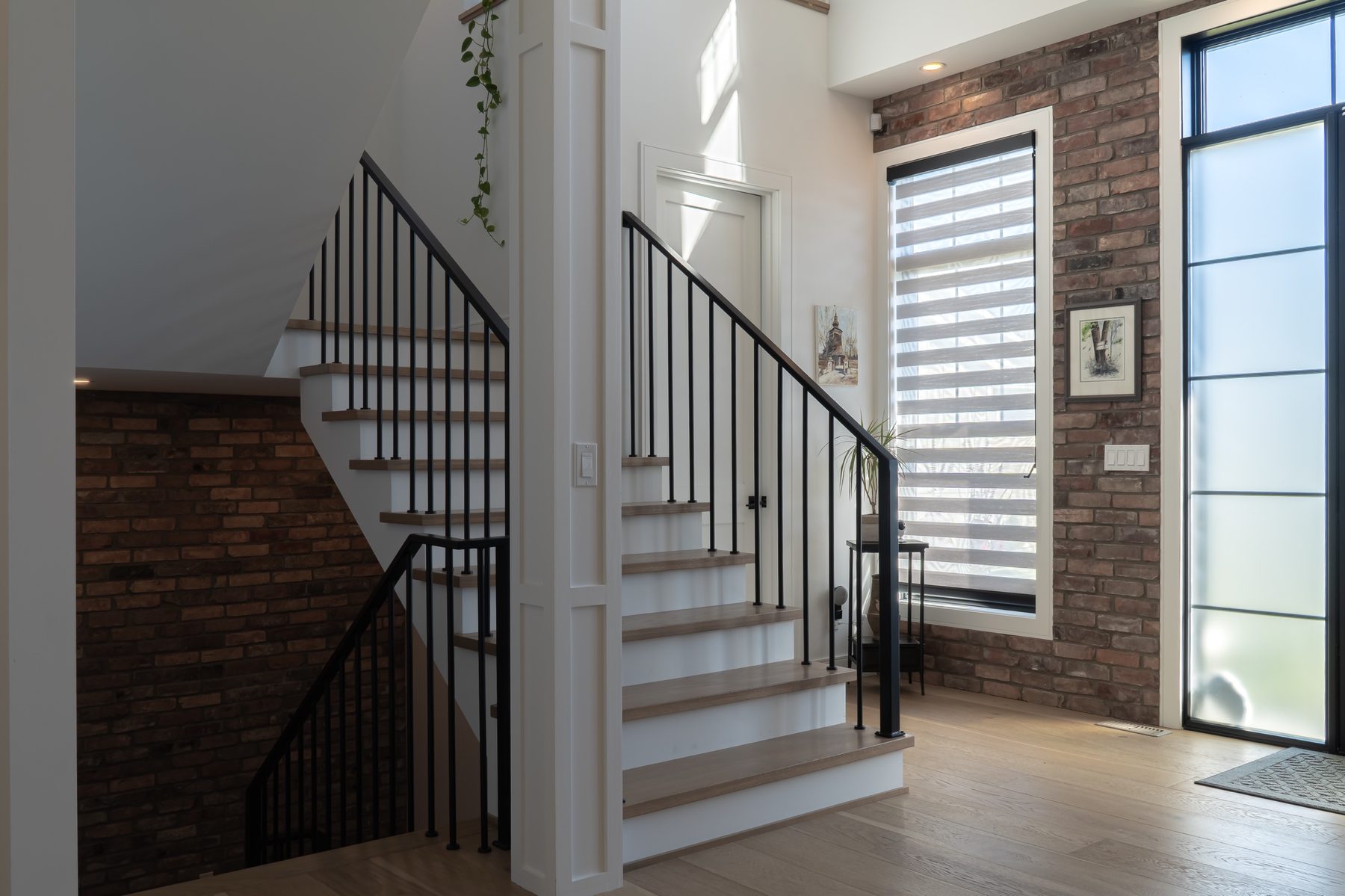 Entry with an oak staircase, black iron balusters and a reclaimed brick wall beside a tall window