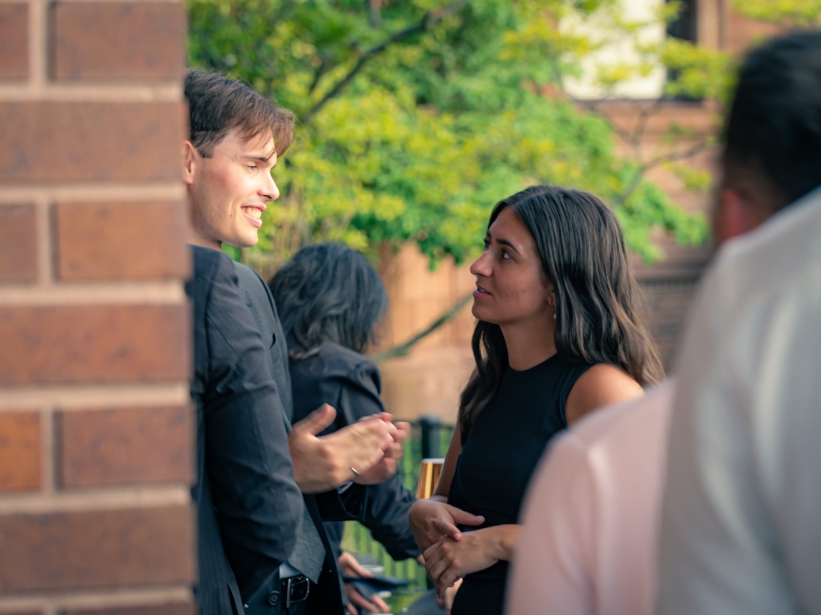 Guests talking at an outdoor evening event beside a brick wall, a man laughing as a woman in black listens