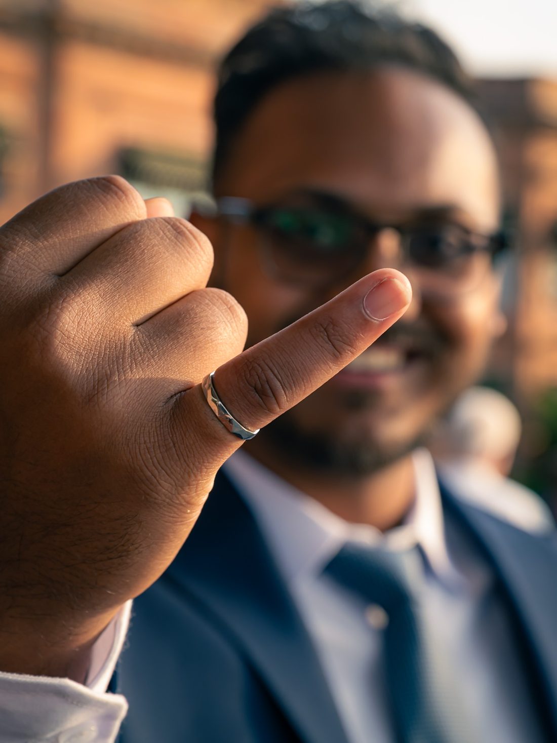 Close-up of a raised hand wearing a silver ring, with a smiling man in a blue suit softly blurred behind