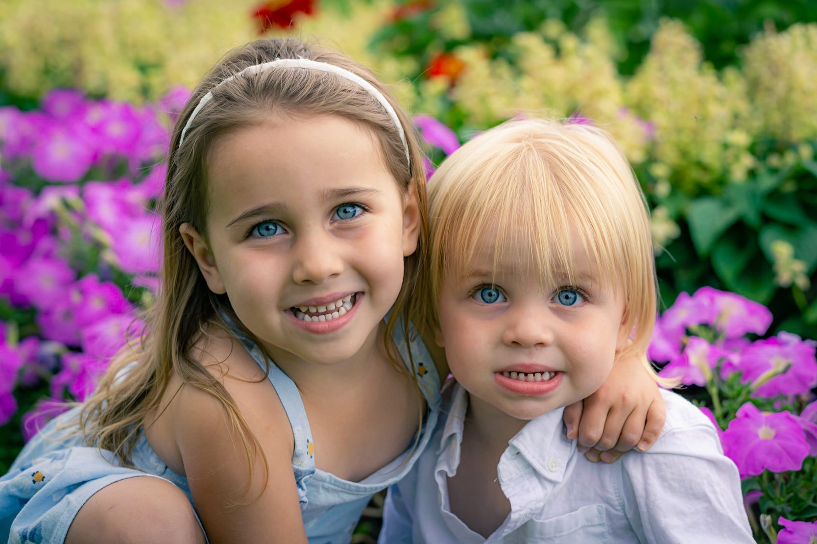 Two young children smiling side by side in front of pink petunias and garden flowers