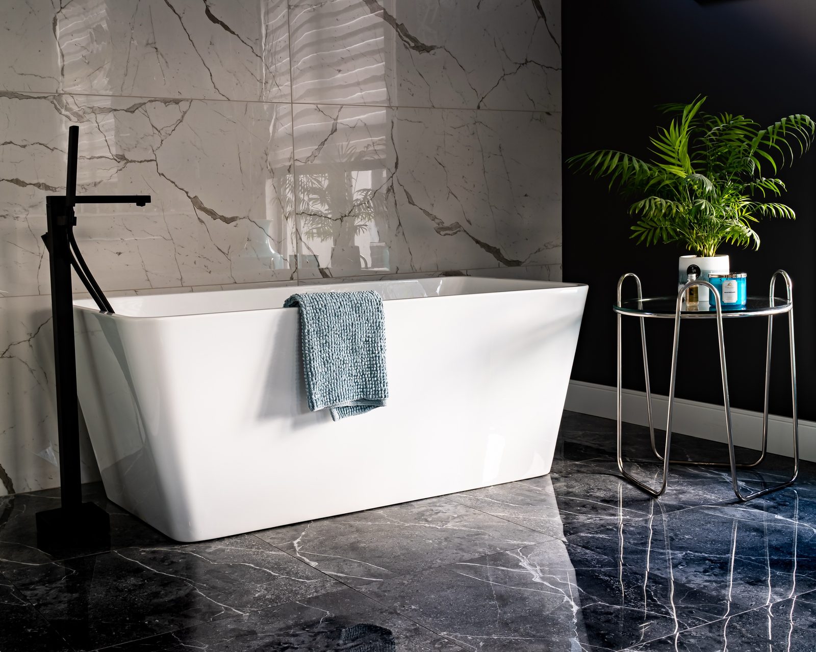 Freestanding white bathtub against a large-format marble wall and dark stone floor, with a black floor-mounted tap and a chrome side table