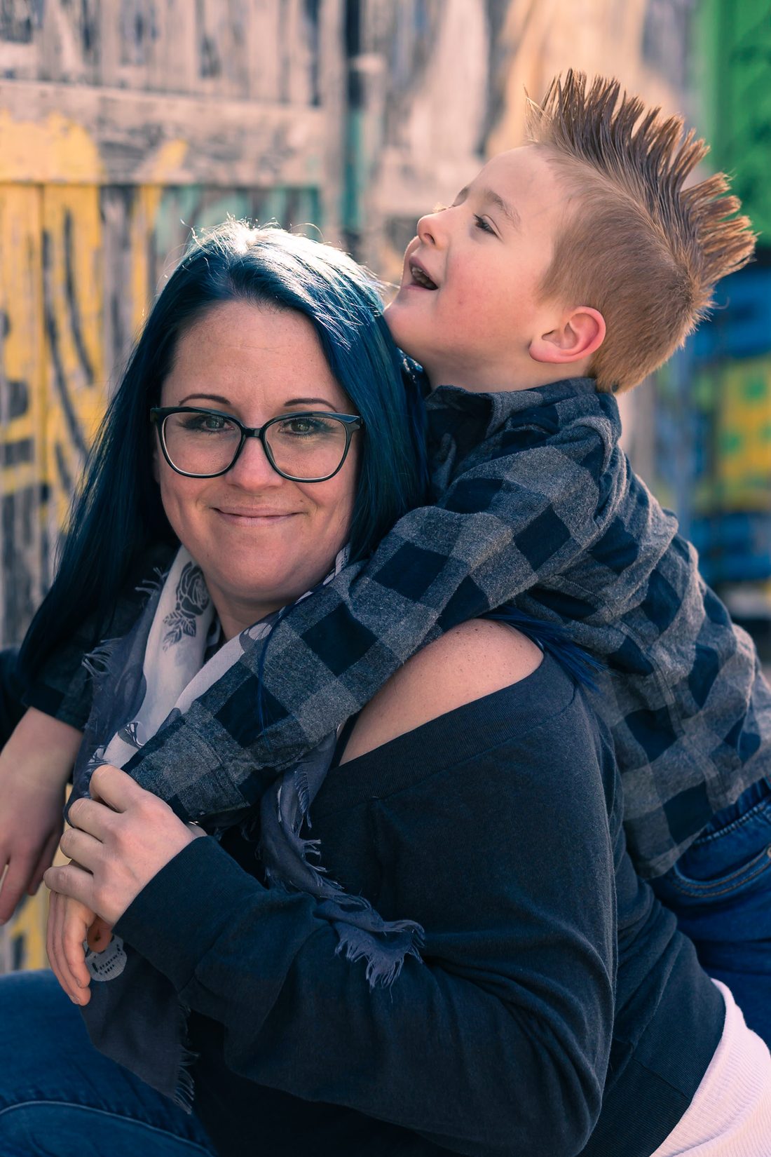 Woman with blue hair and glasses smiling at the camera while her young son hugs her from behind, in front of a graffiti wall
