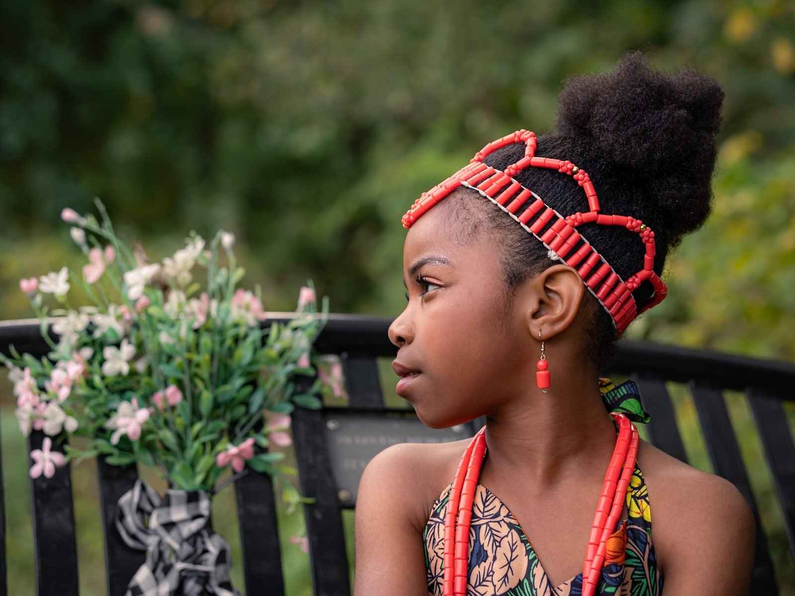 Girl in profile wearing a red beaded crown, matching necklace and earring, beside a bench with flowers