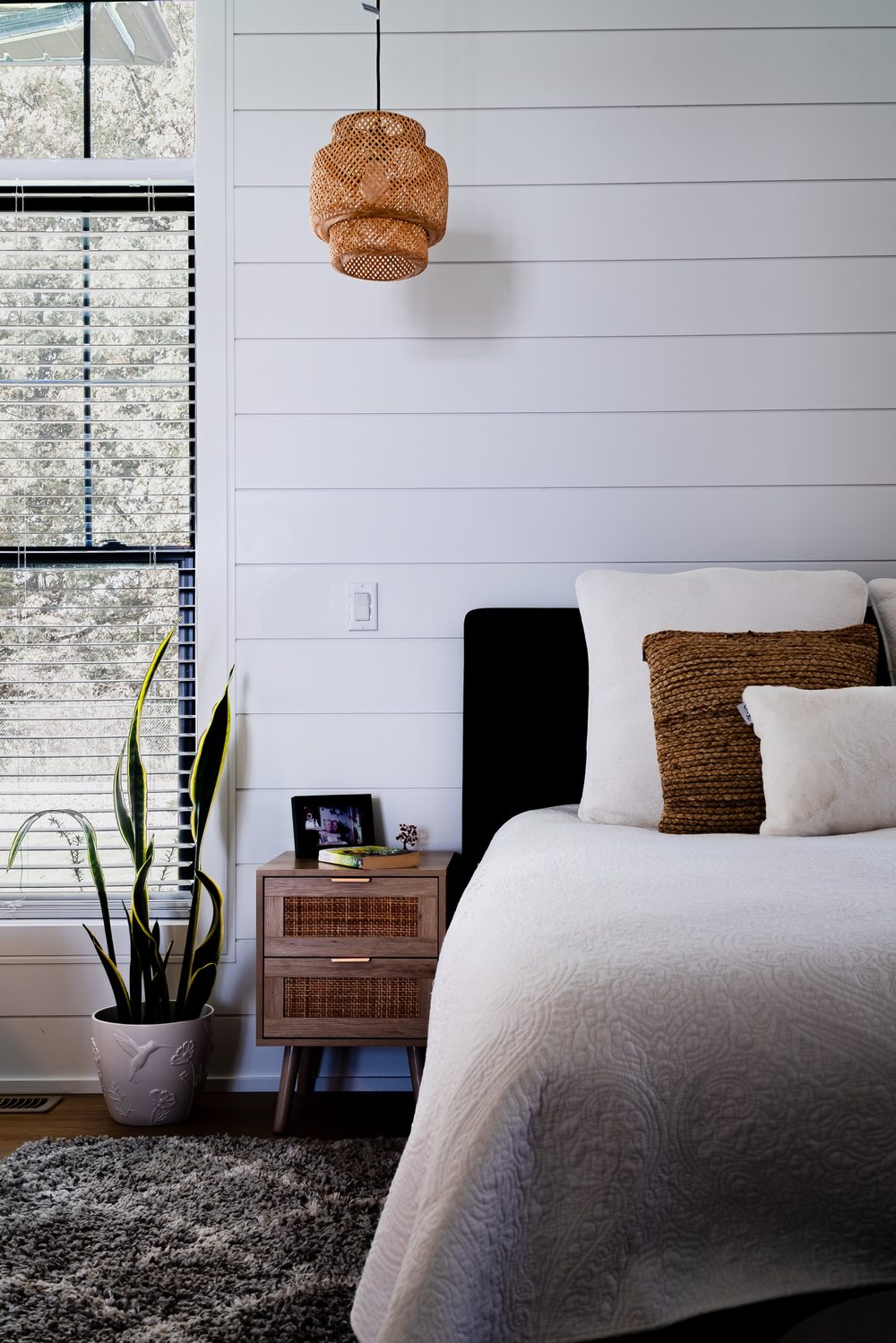 Bedroom corner with a shiplap wall, a woven pendant light, a rattan nightstand and a snake plant by the window