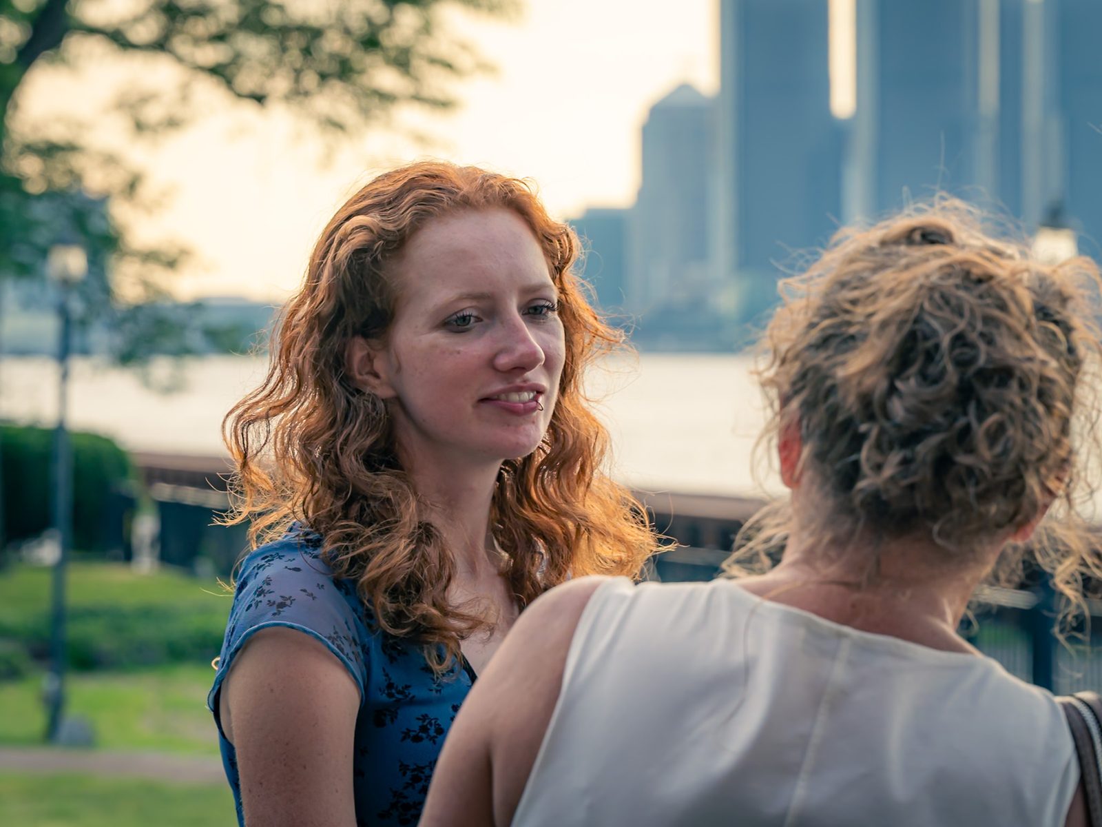 Woman with curly red hair smiling in golden light beside a waterfront, a city skyline behind her